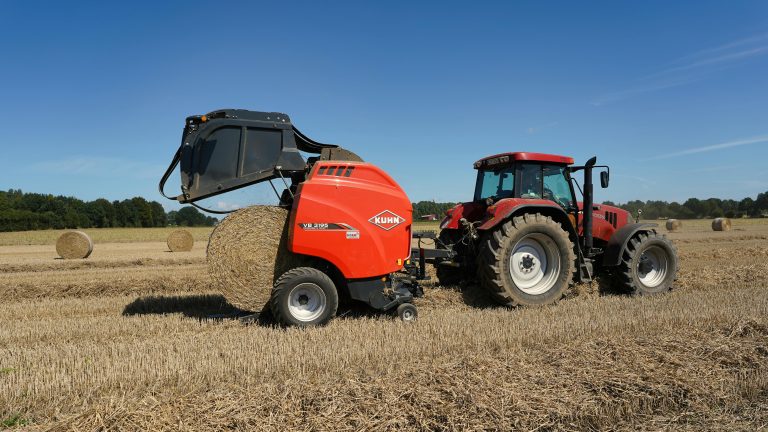 A red tractor and farming machine work in a vast wheat field under a clear blue sky in Hohenhorn, Germany.